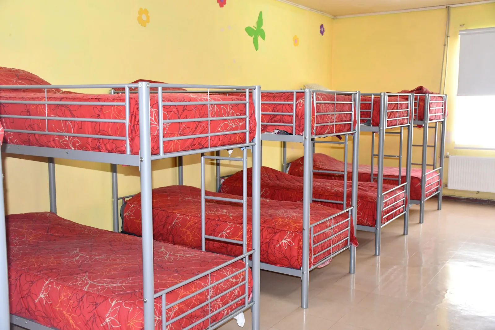 Dormitorio del internado: literas metálicas de dos pisos con colchones rojos, ordenadas contra un muro amarillo con ventanal.