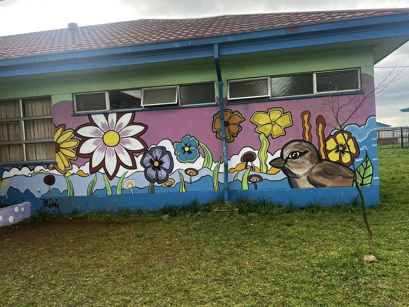 Mural de flores de colores y un chucao de gran tamaño pintado sobre el muro de un pabellón, con césped en primer plano.