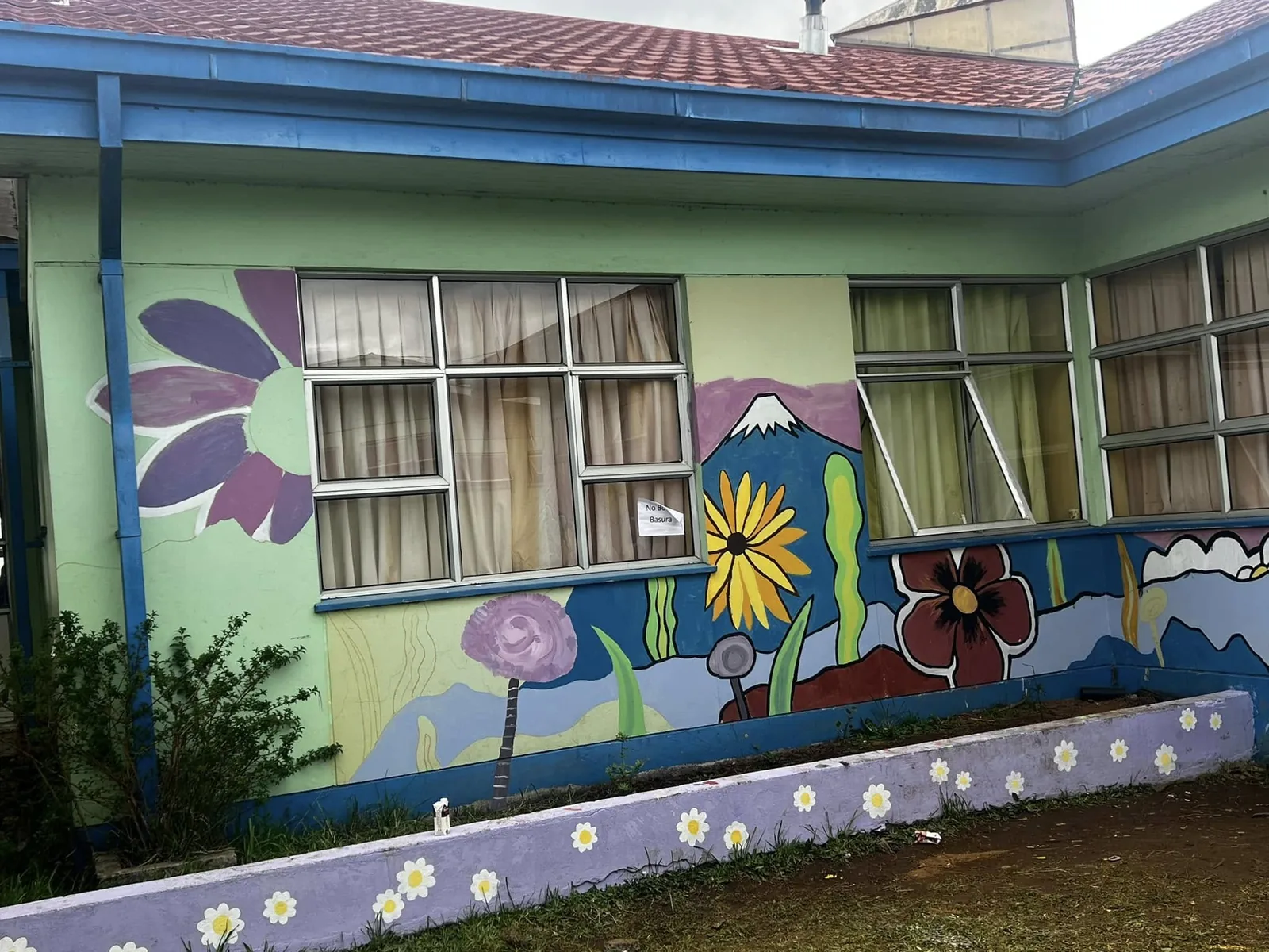 Esquina de un pabellón con muros verdes y rosados pintados con flores, montañas y un zócalo azul con margaritas.