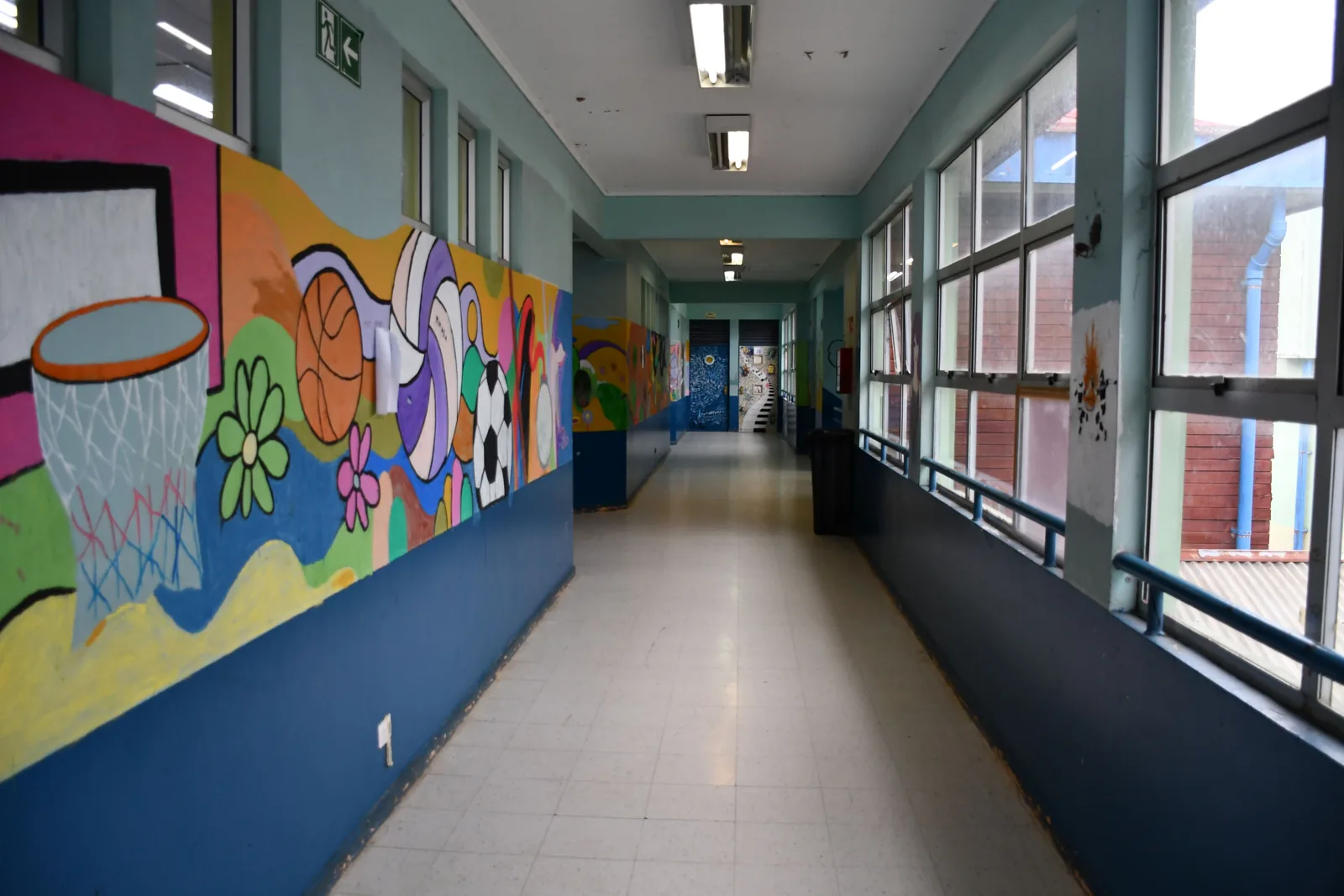 Pasillo interior del liceo con ventanales a un costado y un mural deportivo pintado en el muro contrario.